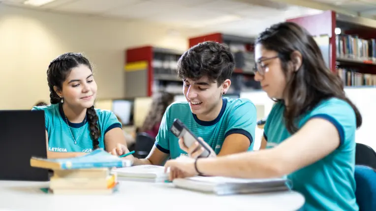três alunos do Bernoulli estudando juntos com cadernos e materiais escolares na biblioteca da escola